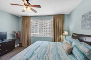 A bedroom with a bed, a ceiling fan, a window with curtains, a TV, and a wall sign. - Shores of Panama by iTrip Panama City Beach (Panama City Beach)