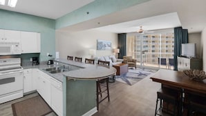 A modern kitchen with white cabinets, a microwave, and a coffee maker. There is a dining area with a round table and chairs, and a living room with a sofa and a TV. - Shores of Panama by iTrip Panama City Beach (Panama City Beach)