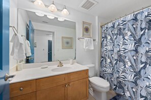 Un bagno con un lavandino bianco, un mobile bagno in legno e un water. C'è una tenda da doccia con un motivo a foglie e un quadro appeso al muro.