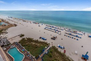 A beach with rows of sun loungers and umbrellas, a swimming pool, and a lifeguard tower.