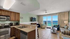 A modern kitchen with a dishwasher, microwave, and stove. There is a dining area with a table and chairs, and a living room with a sofa and a view of the ocean.