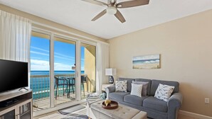 A living room with a TV, a sofa, a coffee table, and a balcony with a view of the ocean.