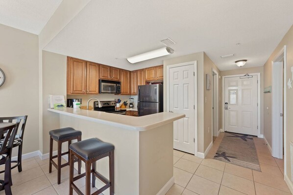 A kitchen with wooden cabinets, a stainless steel refrigerator, a microwave, and a sink. There are three bar stools and a clock on the wall.