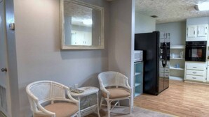 A compact kitchen with white cabinetry, a black refrigerator, and a microwave. Two wicker chairs with cushions are placed in front of a mirror. - It's Five O'clock Somewhere 6/3) (Panama City Beach)