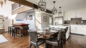 Un comedor con una mesa de madera dispuesta para seis personas, rodeada de sillas de cuero. A la izquierda, se observa una zona de bar con taburetes. La cocina tiene armarios blancos y un salpicadero de ladrillo.