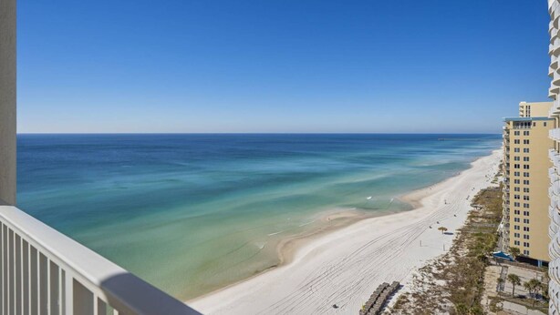 A beach view from a balcony overlooking a sandy shore with clear blue water, adjacent to a multi-story building with balconies. - Ocean Ritz by iTrip Panama City Beach (Panama City Beach)