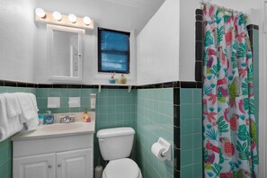Un baño con inodoro, lavabo y tocador. Hay una ventana con persianas, una cortina de ducha con estampado tropical y un espejo con luces arriba.