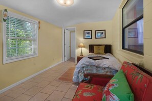 Un dormitorio con una cama, una mesita de noche, una lámpara, una ventana con persianas y una puerta que da a otra habitación.
