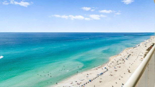 Une plage aux eaux turquoise et limpides, une jetée qui s’avance dans la mer, et une rive sablonneuse parsemée de parasols et de quelques personnes.