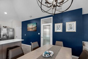 Une cuisine moderne avec un réfrigérateur en acier inoxydable, un coin repas avec une table et des chaises en bois, et un mur d’accent bleu orné de deux tableaux encadrés.