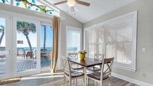 Un espace repas comprenant une table et des chaises, de grandes fenêtres équipées de stores, un ventilateur de plafond et une vue sur la plage.