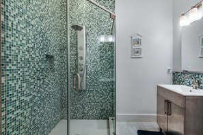 A bathroom with a glass shower enclosure, a sink with a modern faucet, and a tiled wall with a green mosaic pattern. - Parrotdise on the Gulf (Panama City Beach)