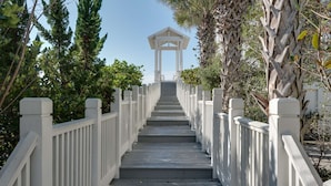En trätrappa leder upp till en vit pergola, som omges av frodig grönska och palmer.