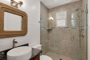 A modern bathroom with a round sink, a toilet, a glass-enclosed shower, and a large mirror with a rope frame.