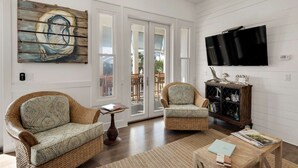 Un salon comprenant deux fauteuils en osier, une table basse, un téléviseur fixé au mur et un meuble en bois orné d’objets décoratifs.