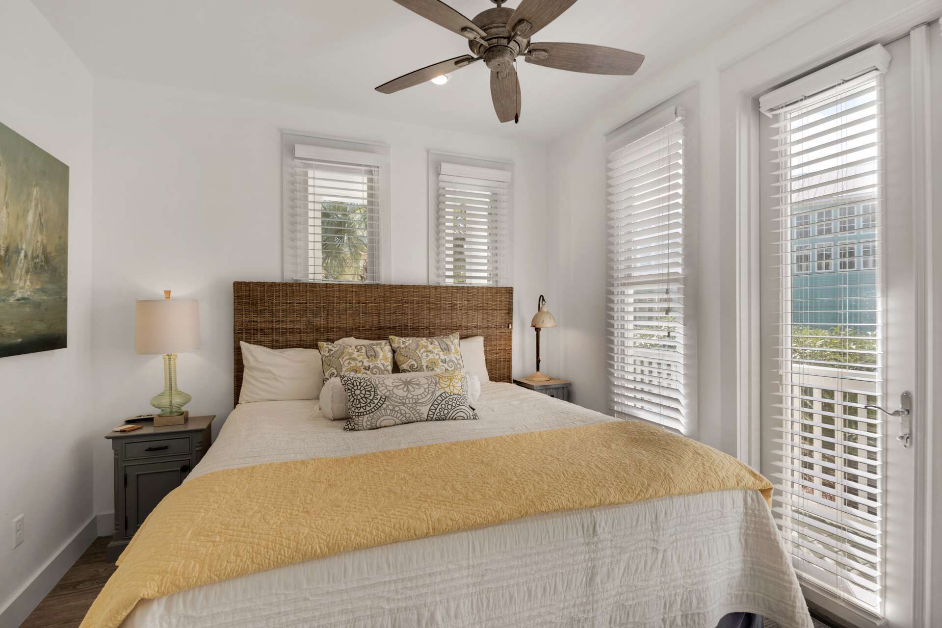 A bedroom with a large bed, a ceiling fan, and a view through the window blinds.