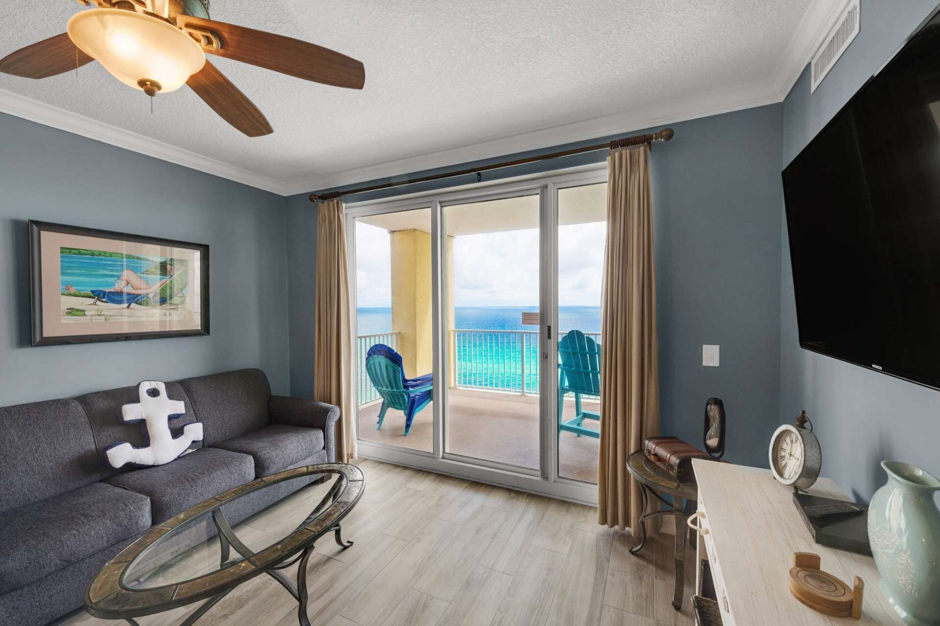 A room with a gray sofa, a glass coffee table, a ceiling fan, a framed picture, a TV, and a balcony with chairs and a view of the ocean.