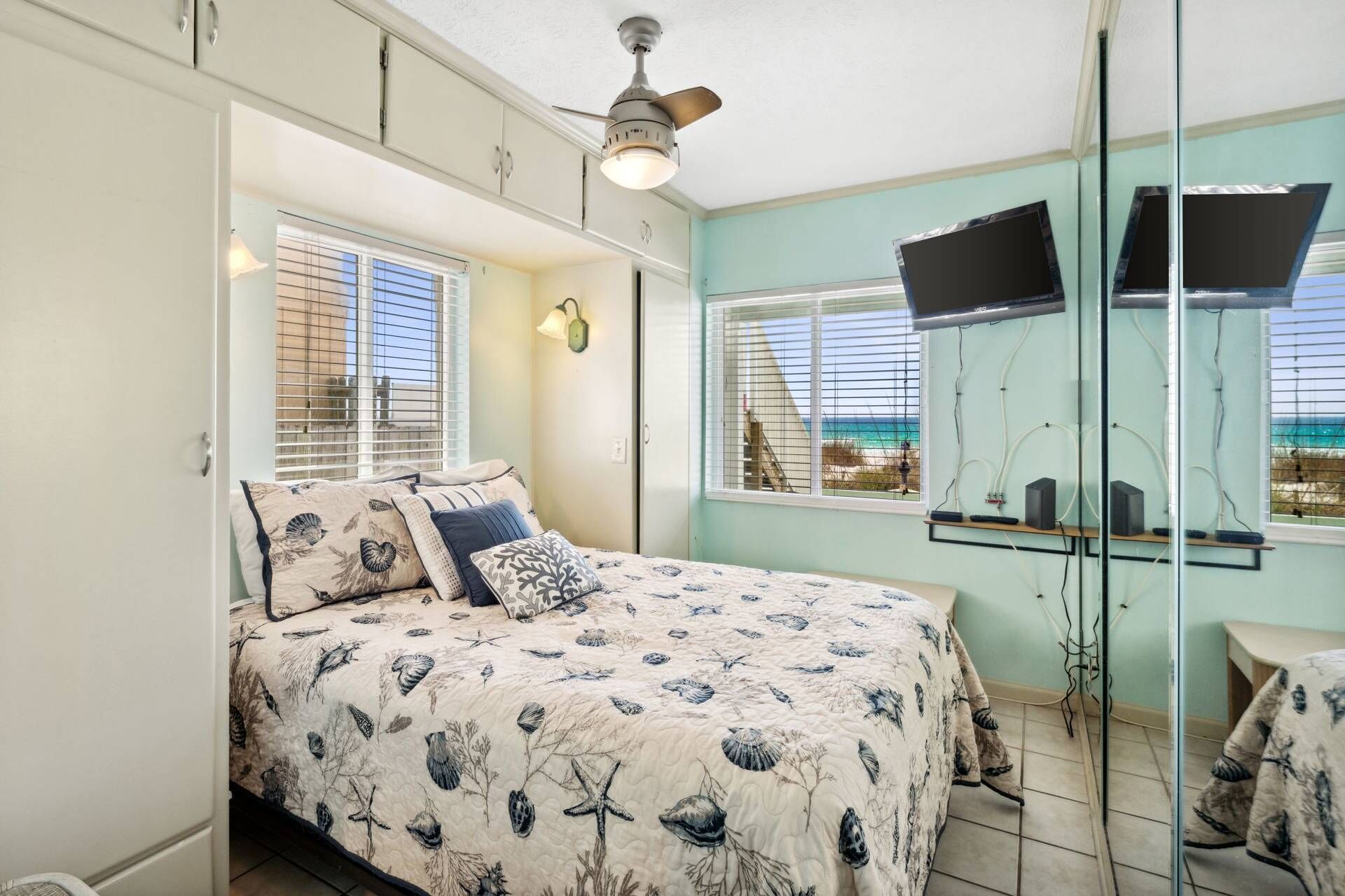 A bedroom with a bed, a ceiling fan, a TV, and a view of the beach.