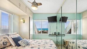Un dormitorio con cama, ventilador de techo, televisor montado en la pared y ventana con vista a la playa.