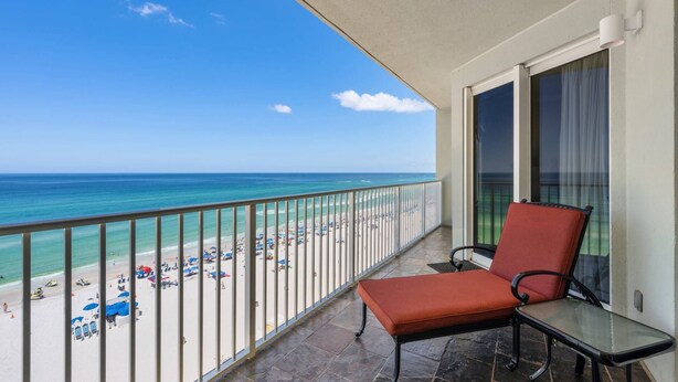A balcony with a red chair and a glass table, overlooking a beach with umbrellas and people. - Marisol by iTrip Panama City Beach (Panama City Beach)