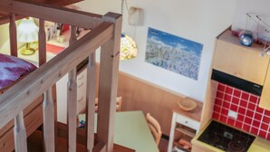 Un escalier en bois mène à une pièce où se trouvent une table verte, une chaise, un poêle et un mur recouvert de carreaux rouges.