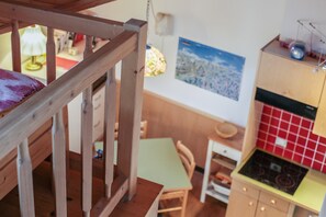 Un escalier en bois mène à une pièce où se trouvent une table verte, une chaise, un poêle et un mur recouvert de carreaux rouges.