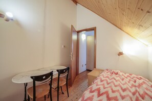 Una habitación con una cama, una mesita, dos sillas, techo de madera y una puerta que da a otra habitación.