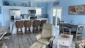 Una cocina moderna con armarios blancos y una isla central, una zona de comedor con mesa y sillas de madera, y un espacio de descanso con una silla acolchada y reposapiés.