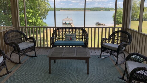 A covered outdoor seating area with wicker chairs and a table, overlooking a lake with a dock and boat. - THE Premier Clemson /Seneca Lake Home  (Seneca)