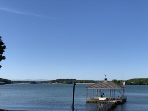 Um cais com um pavilhão coberto em um lago calmo, rodeado por árvores e colinas ao longe.
