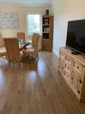 Un comedor con sillas de mimbre, una mesa de vidrio, un mueble de madera con cajones, un televisor y una estantería con un recorte en forma de corazón.