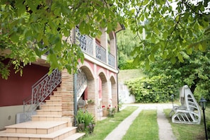 Ein Gebäude mit einer Treppe, die zu einem Balkon führt, umgeben von Grünpflanzen und einem Weg.