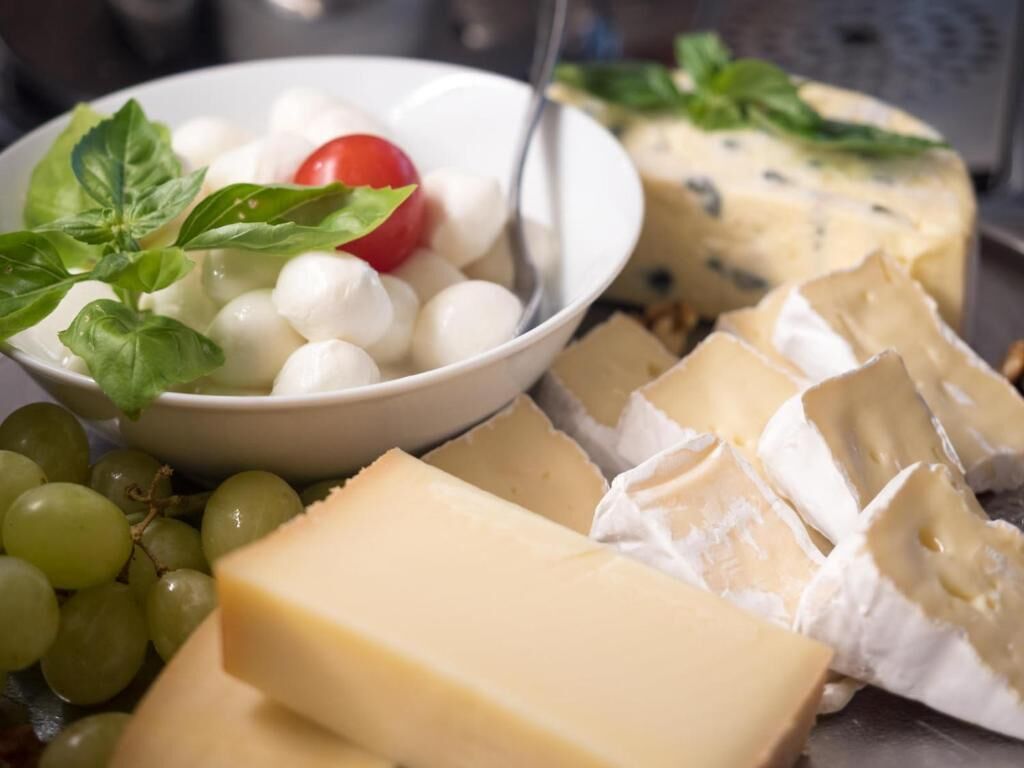 Eine Auswahl an Käsesorten, darunter Mozzarella-Kugeln, ein Käselaib und eine Scheibe Blauschimmelkäse, begleitet von Trauben und einer Kirschtomate.
