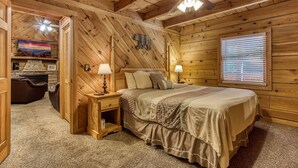 A cozy log cabin bedroom with a bed, two bedside tables with lamps, a fireplace, a television, and a window with blinds.