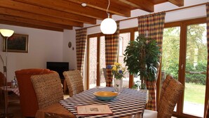 A cozy living room with a wooden ceiling, a dining table with a checkered tablecloth, wicker chairs, and a large window with plaid curtains. - Lütt Hüs, Deutschland (Grünendeich)