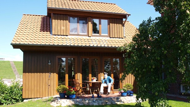 A wooden house with a tiled roof, a porch with a table and chairs, and a garden with potted plants. - Lütt Hüs, Deutschland (Grünendeich)