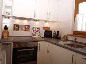 A modern kitchen with white cabinets, a microwave, a coffee maker, and a toaster. There is a window with a curtain and a sink with a faucet. - Lütt Hüs, Deutschland (Grünendeich)