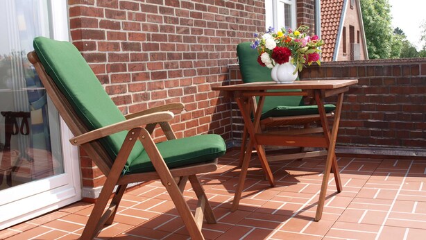 A patio with two wooden chairs and a table, a white vase with flowers, and a brick wall. - Ferienwohung Bootsdeck - Villa Elbdeich (Grünendeich)