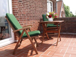 Une terrasse avec deux chaises en bois et une table, un vase blanc rempli de fleurs et un mur en briques.