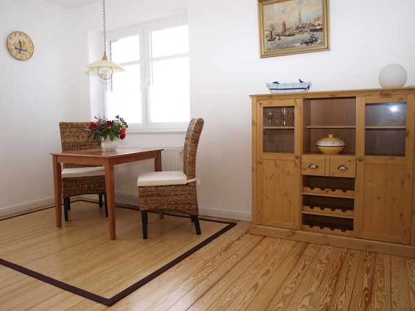 Un coin repas avec une table et des chaises en bois, un vase de fleurs, un buffet en bois contenant des verres à vin et une horloge au mur.