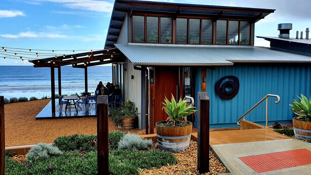 A coastal building with a deck overlooking the sea, featuring a wooden pergola, outdoor seating, and potted plants. - Luxury beach stay,  close to the famous SA wine regions (Aldinga)