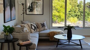 A modern living room with a sofa, a coffee table, and a large window offering a view of greenery. - Luxury beach stay,  close to the famous SA wine regions (Aldinga)