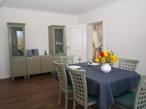 Ein Essbereich mit Tisch, gedeckt für vier Personen, umgeben von Stühlen. Links befindet sich ein Schrank mit Glastüren, und eine weiße Tür führt in einen weiteren Raum. Auf dem Tisch steht eine Vase mit gelben Blumen.