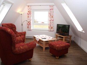 A cozy living room with a red armchair, ottoman, and a wooden coffee table. A television is on a stand, and there's a window with curtains. - Ferienwohnung Oberland - Haus Helgoland (Grünendeich)