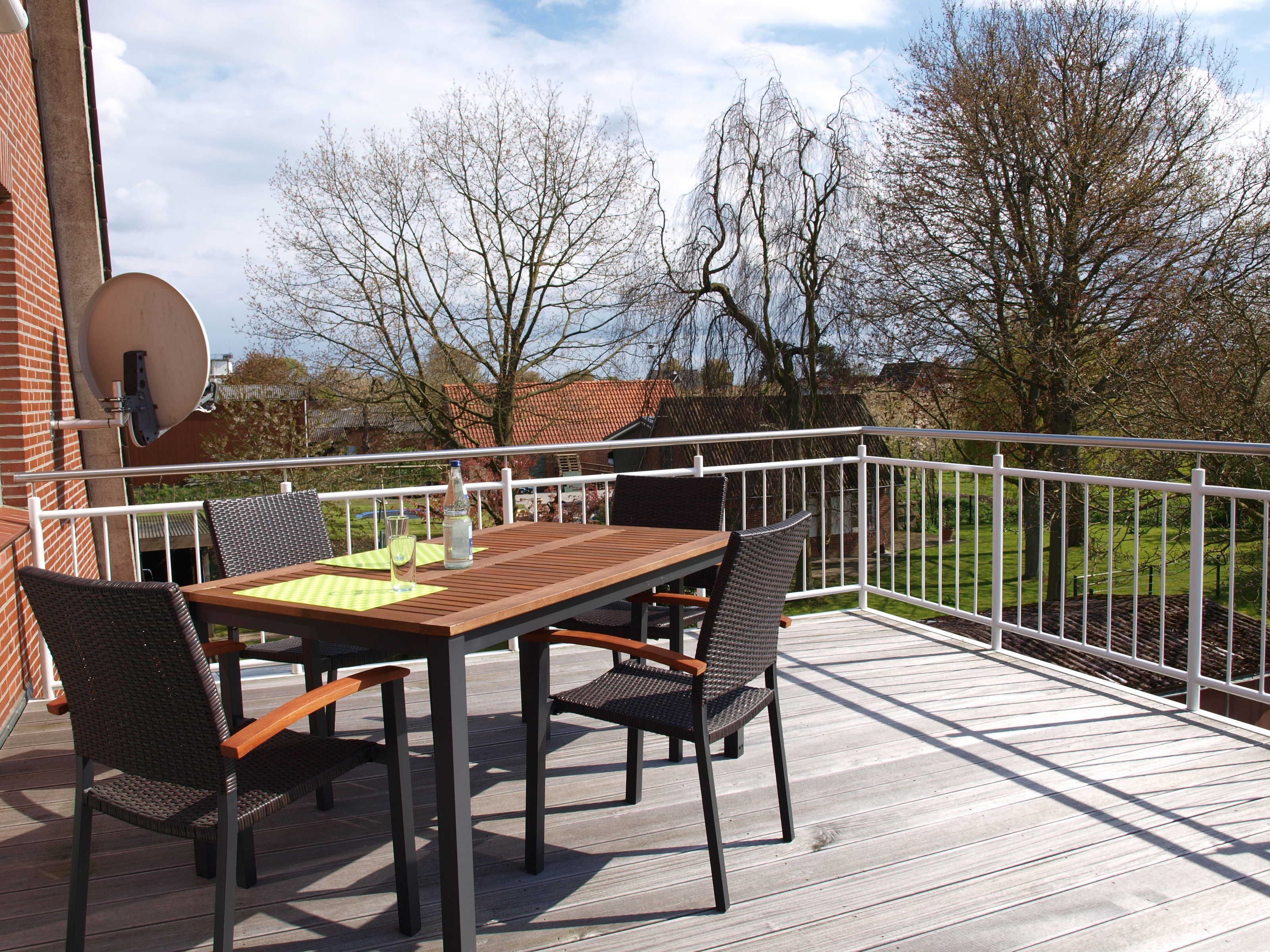 Een houten terras met een tafel en stoelen, een witte reling, en een uitzicht op een groen landschap met bomen en een huis op de achtergrond.