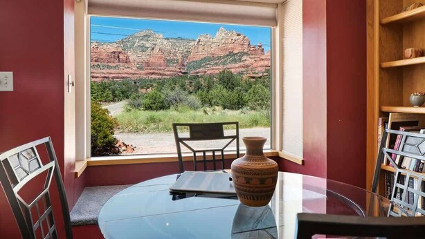 A room with a large window offering a view of red rock formations, a round glass table, a chair, and a bookshelf. - Ranch Style Home Uptown Sedona, View/BBQ/Patio! (Sedona)
