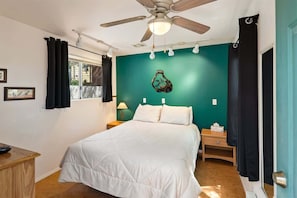 Une chambre à coucher comprenant un lit, un ventilateur de plafond, une fenêtre avec des rideaux, une table de chevet avec une lampe et un vase fixé au mur.