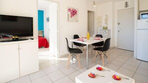 A modern kitchen with white cabinets, a dining area with a white table and black chairs, a flat-screen TV on a white cabinet, and a door with a symbol. - Le Grand Galion by Interhome (Port Camargue)