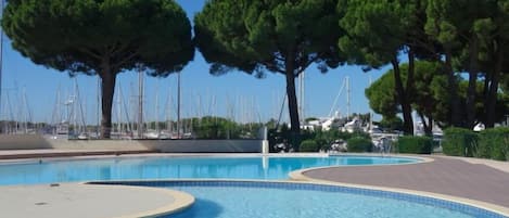 Una piscina de borde curvo, rodeada de árboles y un puerto deportivo con barcos al fondo.