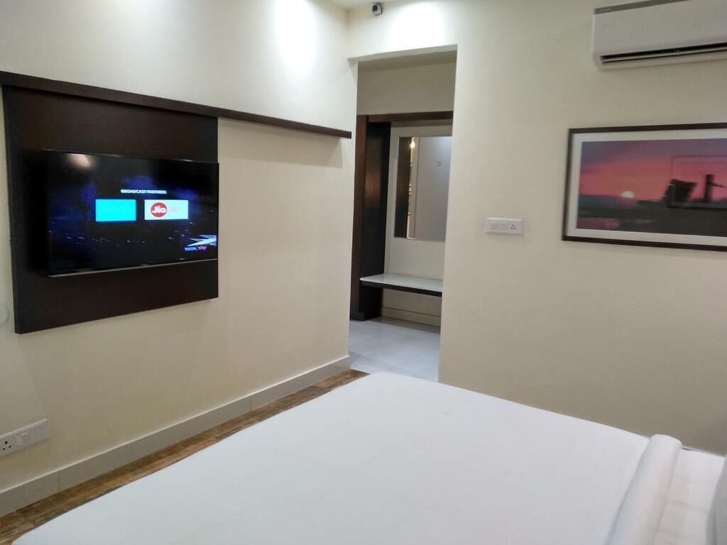 A hotel room with a flat-screen TV mounted on the wall, a bed with white bedding, a framed picture on the wall, and an air conditioning unit.
